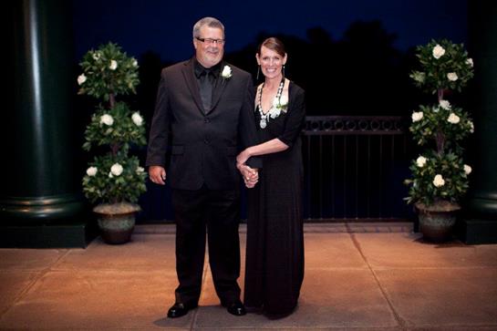 Couple stands hand in hand, dressed elegantly, ready to celebrate a meaningful event outdoors.