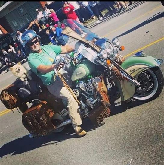 A man proudly rides his motorcycle with a dog comfortably seated beside him at a local event.