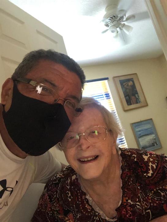 A man wearing a mask smiles beside an elderly woman, both enjoying a warm moment together indoors.