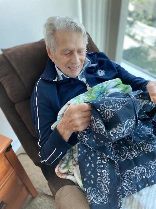 An elderly man smiles as he knits a colorful blanket in a cozy indoor setting.