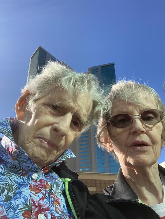 Two older people are taking a selfie together with modern buildings in the background.