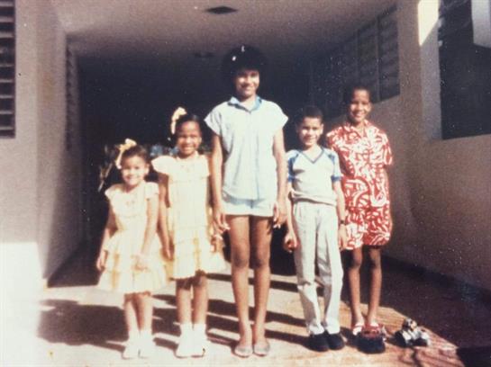Five children stand together smiling in casual outfits at a school entrance on a bright day.
