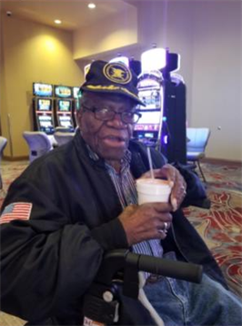 Elderly man in glasses and cap sits in a casino with slot machines, sipping a drink.