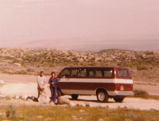 Travelers stand next to an old van on a winding road with hills in the background during daylight.