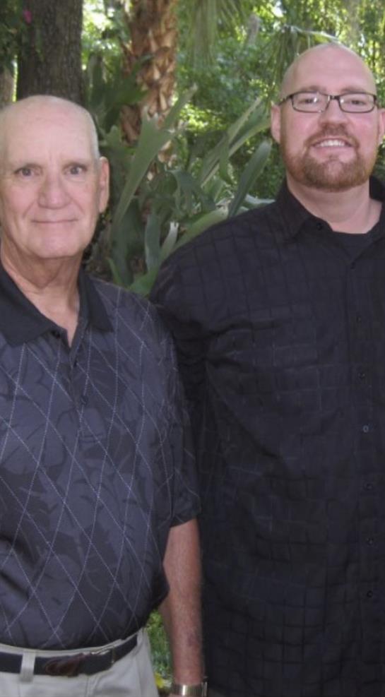 Two smiling men stand side by side amid greenery, enjoying each other's company at a casual event.