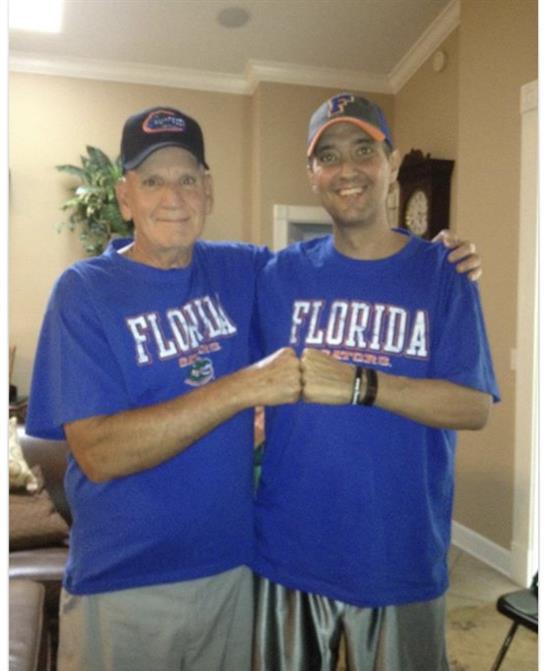 Two men share a joyful moment, wearing matching blue Florida shirts, smiling in a living room.