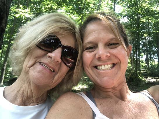 Two women relax in a wooded area, sharing smiles and enjoying each other's company on a sunny day.