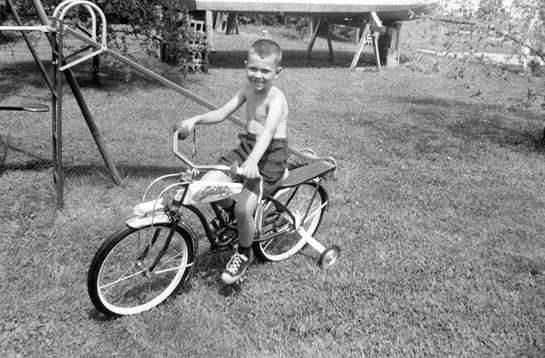 A young boy rides a classic bicycle in a grassy backyard with a swing set and slide.