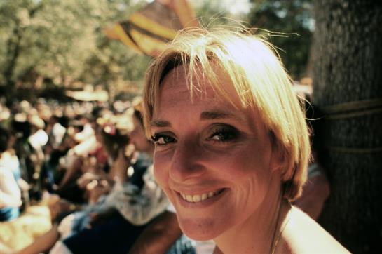Woman with short blonde hair smiles warmly at a lively outdoor festival, surrounded by people.