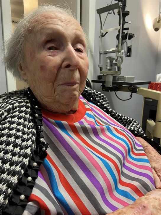 An elderly woman sits in an eye clinic wearing a colorful striped shirt, smiling softly.
