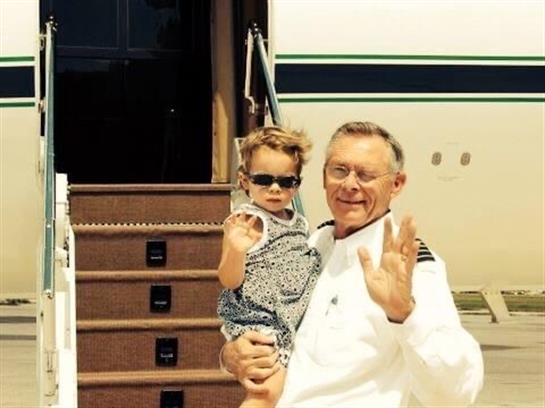 A pilot in uniform holds a child, both smiling and waving with their hands at an airfield.
