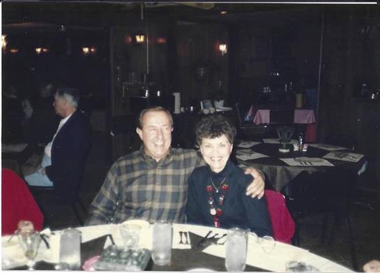Two seniors happily share a table at a bustling restaurant, smiling warmly at each other.