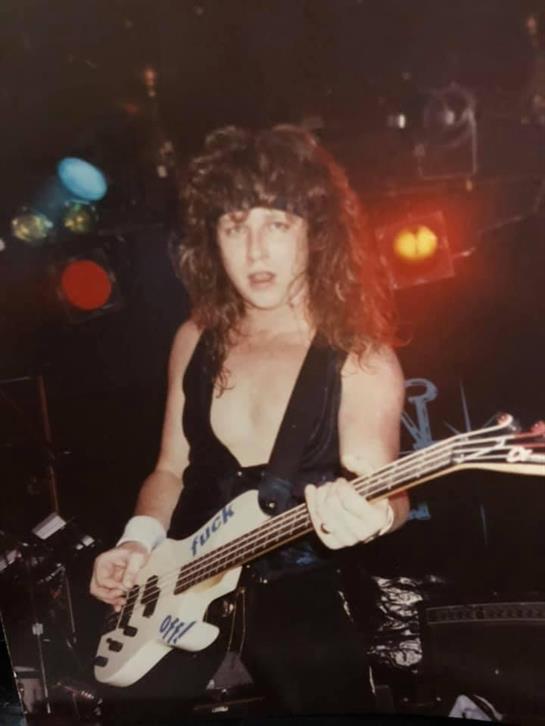 Musician in a black outfit plays bass guitar under colorful stage lights during a performance.