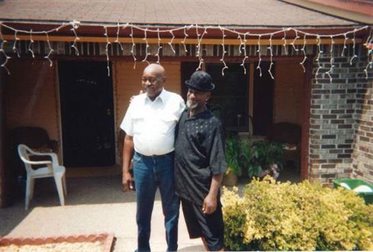 Two men stand closely together in front of a home adorned with festive lights.