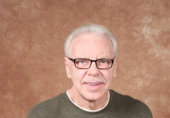 An elderly man with gray hair and glasses smiles at the camera, exuding warmth and friendliness.