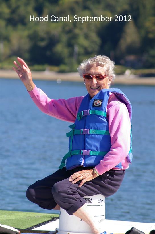 A senior woman in a life jacket kayaks on a serene lake, waving joyfully at bystanders.