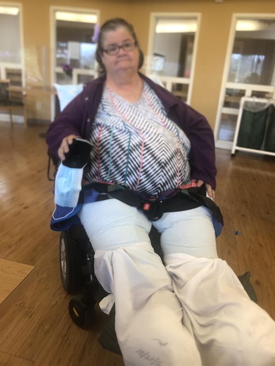 A woman uses a wheelchair and smiles while seated in a community center on a sunny day.