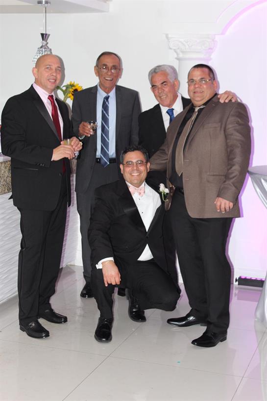 Five well-dressed men pose together cheerfully at a festive event held indoors.
