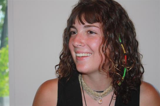 A young woman with curly hair is smiling brightly while wearing layered necklaces at a gathering.