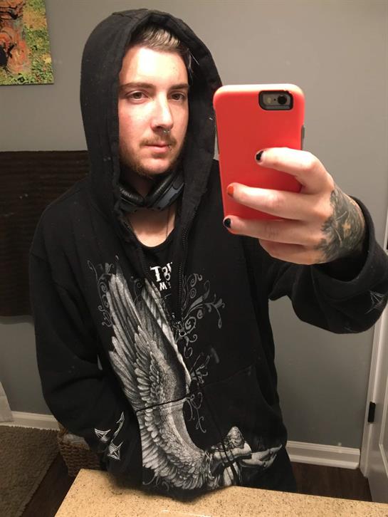 A young man with tattoos poses for a selfie while wearing a hooded sweatshirt in a modern bathroom.