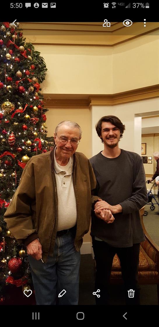Two relatives, an older man and a young adult, share a festive moment by a decorated tree.