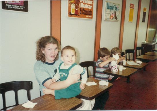 A woman holds a toddler while other children are eating at tables in a warm diner setting.