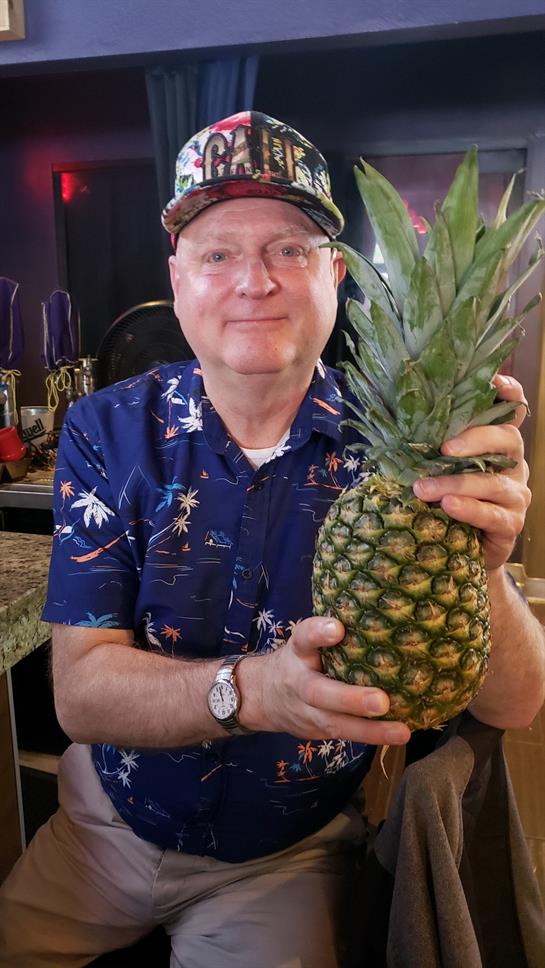 A middle-aged man in a colorful shirt enjoys holding a freshly picked pineapple in his kitchen.