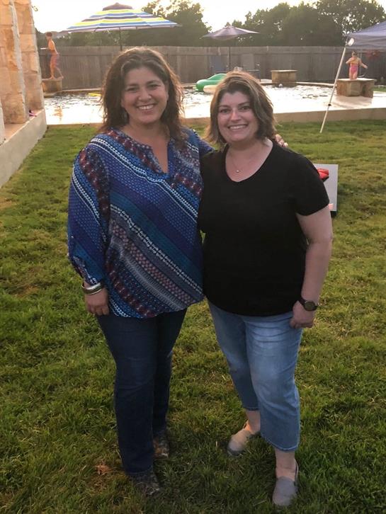 Two women stand together in a grassy backyard, smiling and enjoying their time at a gathering.