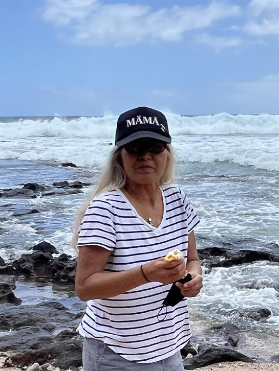 A woman stands near the shore, snacking while waves crash in the background under a blue sky.