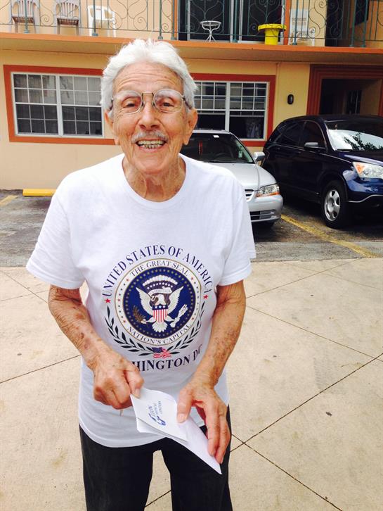 An elderly man is joyfully smiling and holding a piece of paper outside a building with cars nearby.