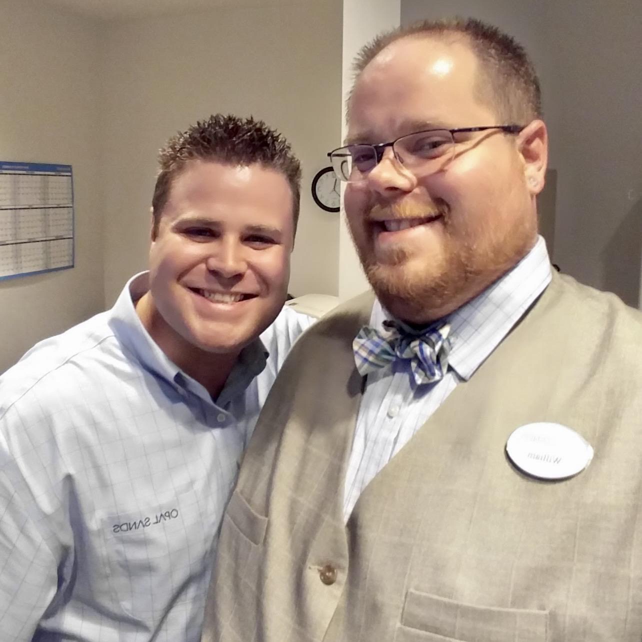 Two men stand close together in an office, both smiling and dressed in professional clothing.