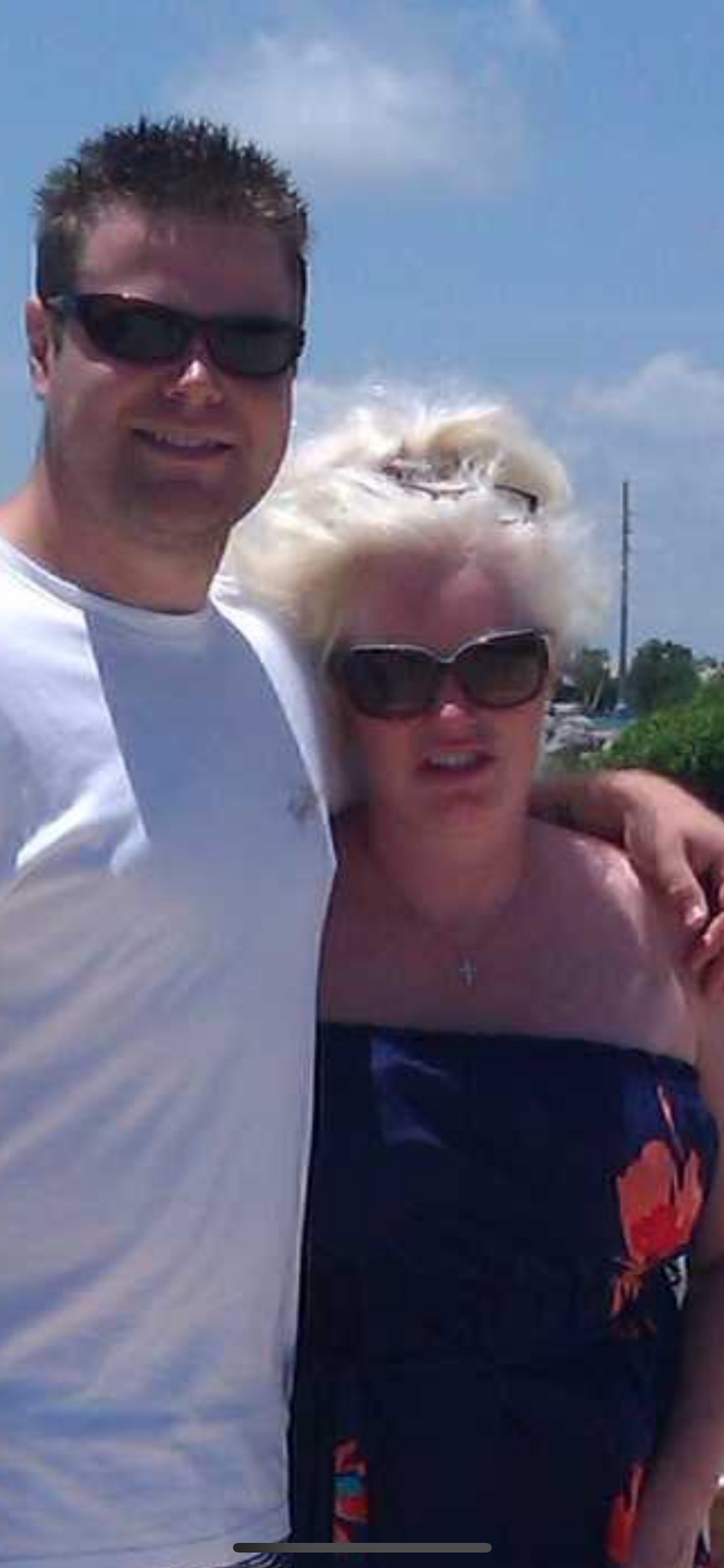Two people stand close together smiling at the beach on a bright sunny day.