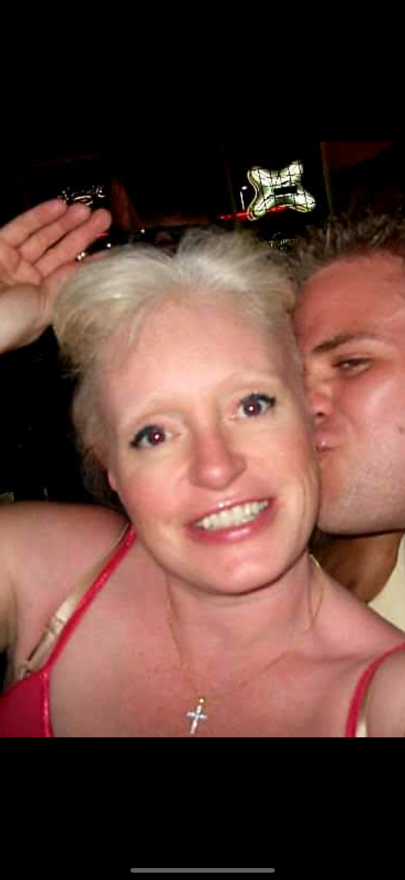 Woman smiles brightly while man kisses her cheek at a festive event with background lights