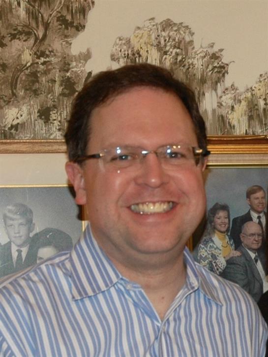 A cheerful man stands happily in front of a wall filled with notable photographs during a gathering.