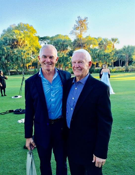 Two men stand together smiling at a lively outdoor gathering surrounded by greenery.