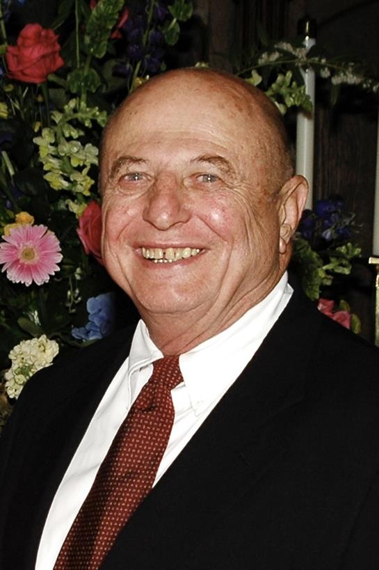 An older man in a suit smiles widely, surrounded by blooming flowers.