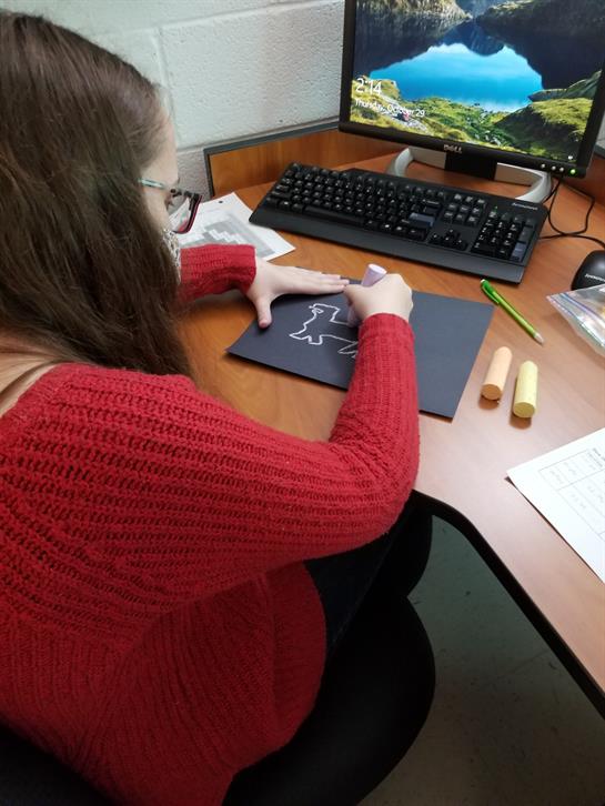 A person is meticulously crafting on dark paper while using tools at a desk with a computer.