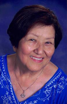 An older woman smiles warmly, dressed in colorful, bright-patterned clothing at a joyful event.