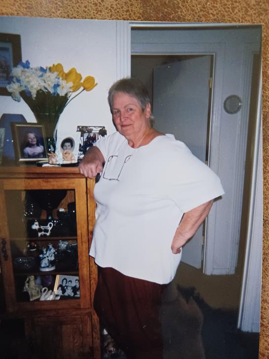 An elderly woman poses with a hand on her hip next to family photos and a floral arrangement.