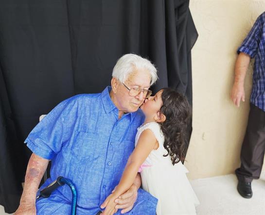 A young girl kisses her grandfather lovingly while sitting in a casual indoor environment.