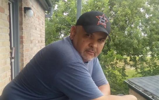A man wearing a cap sits comfortably on a porch, enjoying the sunny weather and greenery.