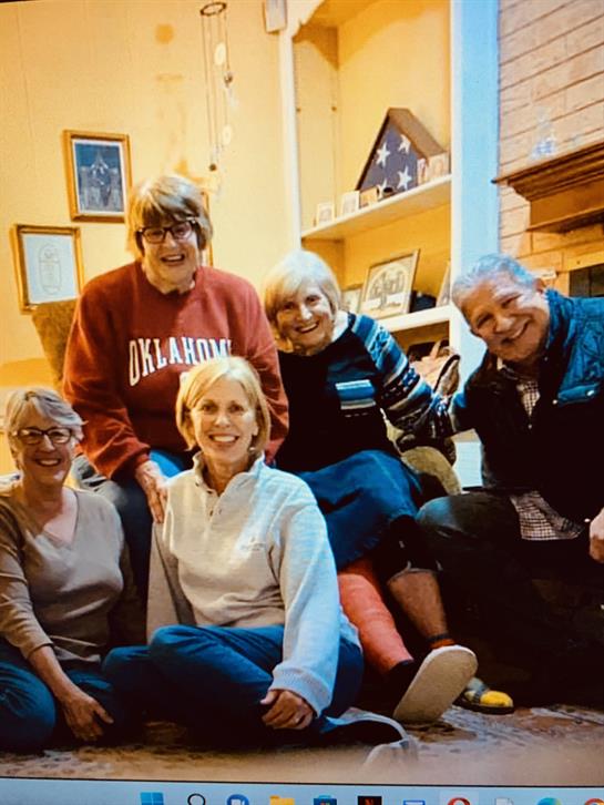 A group of six friends smiles together while relaxing in a warm, inviting living room setting.