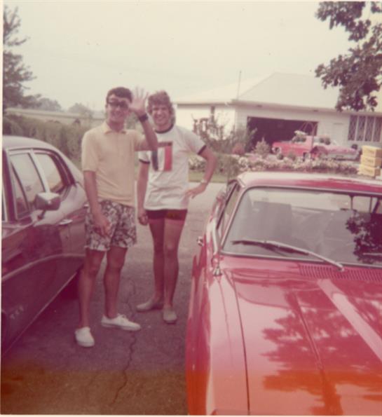 Two young adults stand beside a vintage car, smiling in a suburban area during summer.