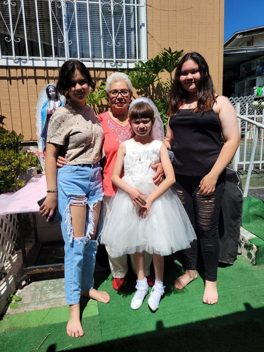 A young girl in a white dress is surrounded by family members, celebrating a special day outdoors.