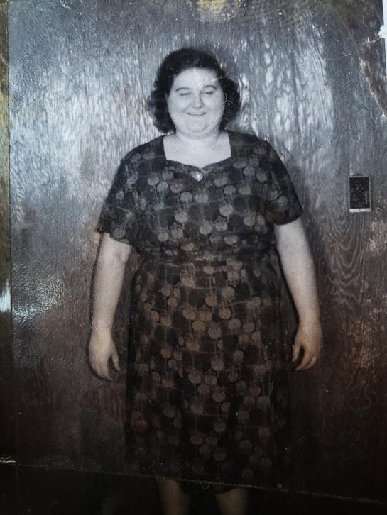 Woman smiles while posing in patterned dress by wooden wall, showcasing vintage fashion.