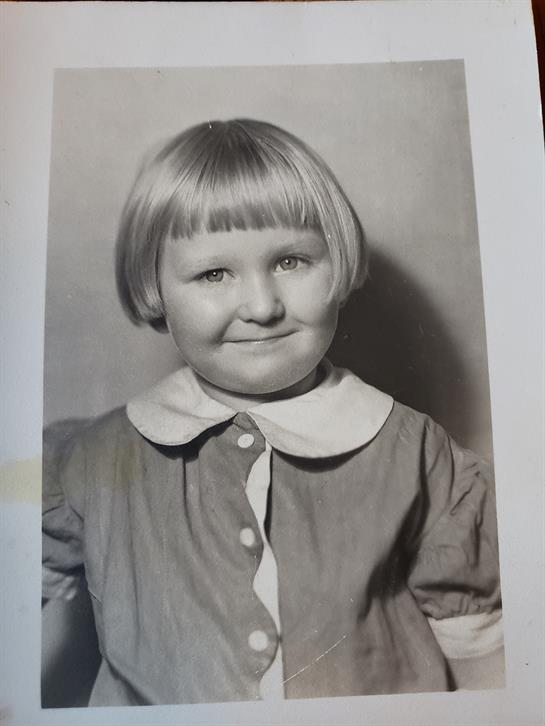 A cheerful young girl with a bob haircut smiles while wearing a collared dress in a studio setting.
