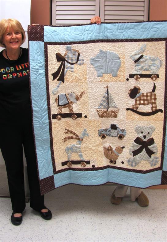 A person displays a colorful quilt with playful animal and toy patterns at a craft event.