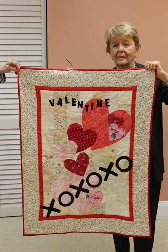 An elderly woman holds up a colorful quilt adorned with heart designs and a loving message.