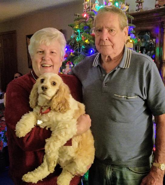 An elderly couple smiles with their dog in a cozy living room decorated for the holidays.