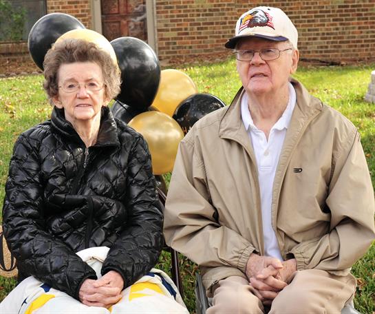 Elderly couple celebrates their anniversary outdoors with black and gold balloons.
